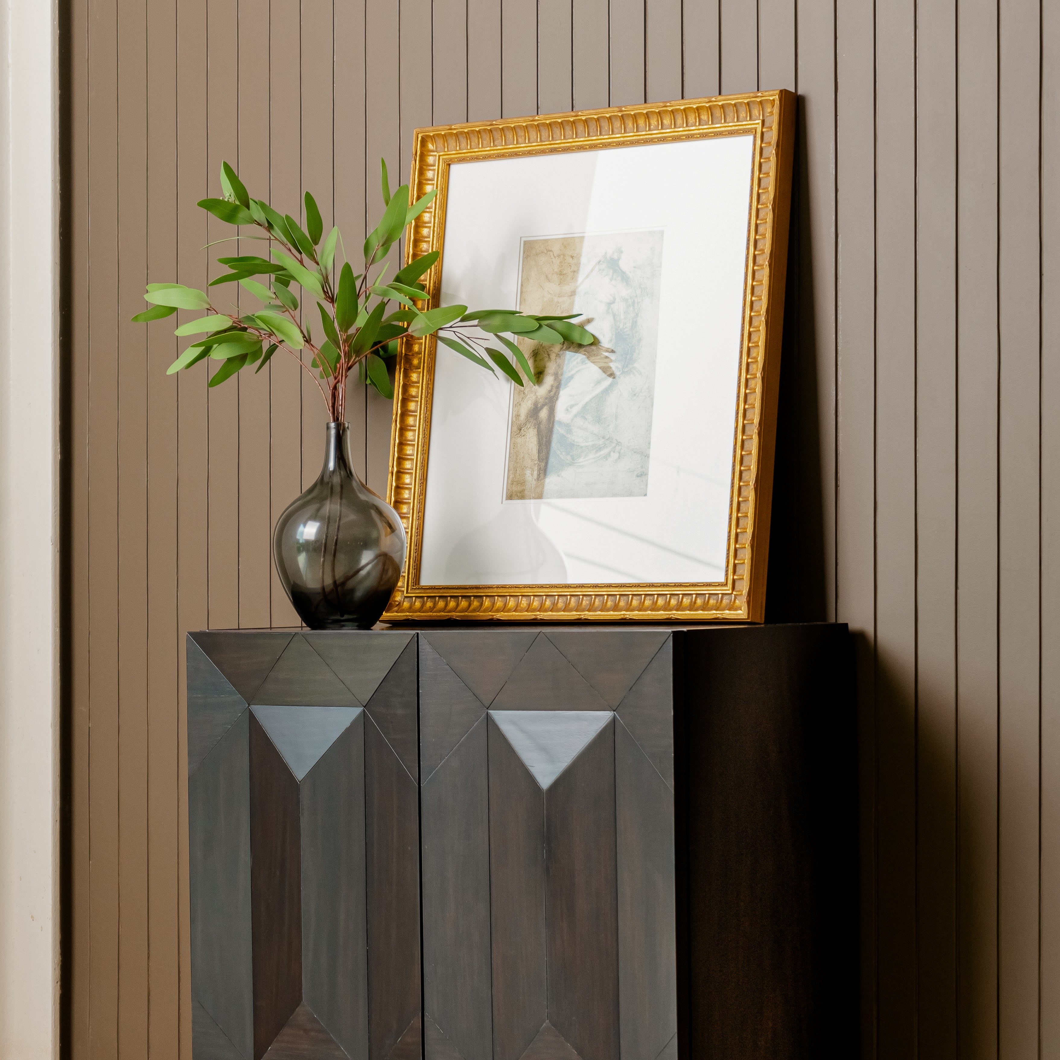 Decorative setup with a gold-framed mirror, vase with greenery, and Black Averi Modular Cabinet against a striped wall.