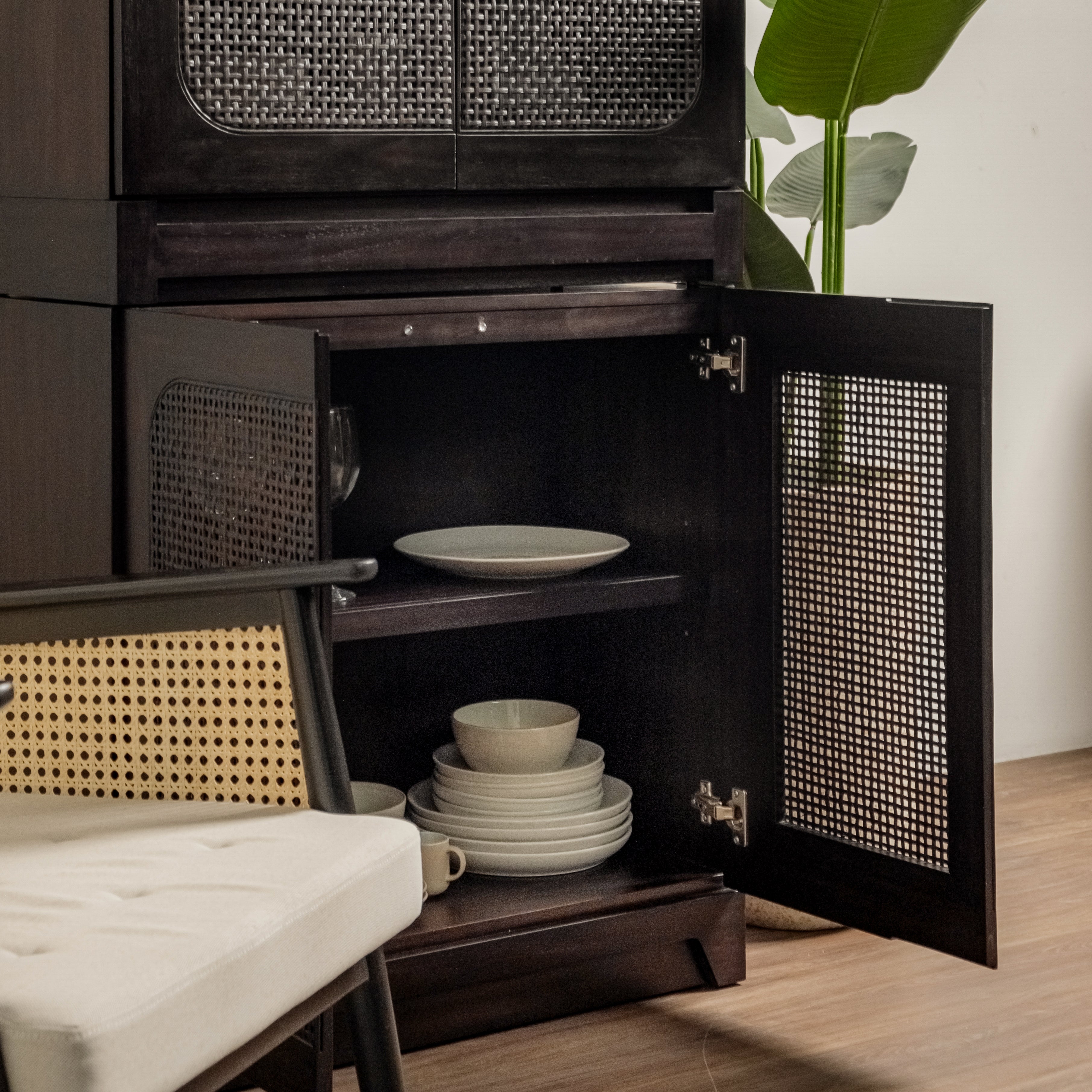 Wooden cabinet with open doors revealing stacked ceramic plates and bowls, with a chair and plant in the foreground.