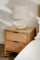 Wooden nightstand with wicker drawers and a white lamp on top against a neutral wall.