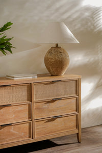 Wooden dresser with wicker drawers and a textured table lamp on top against a neutral wall.