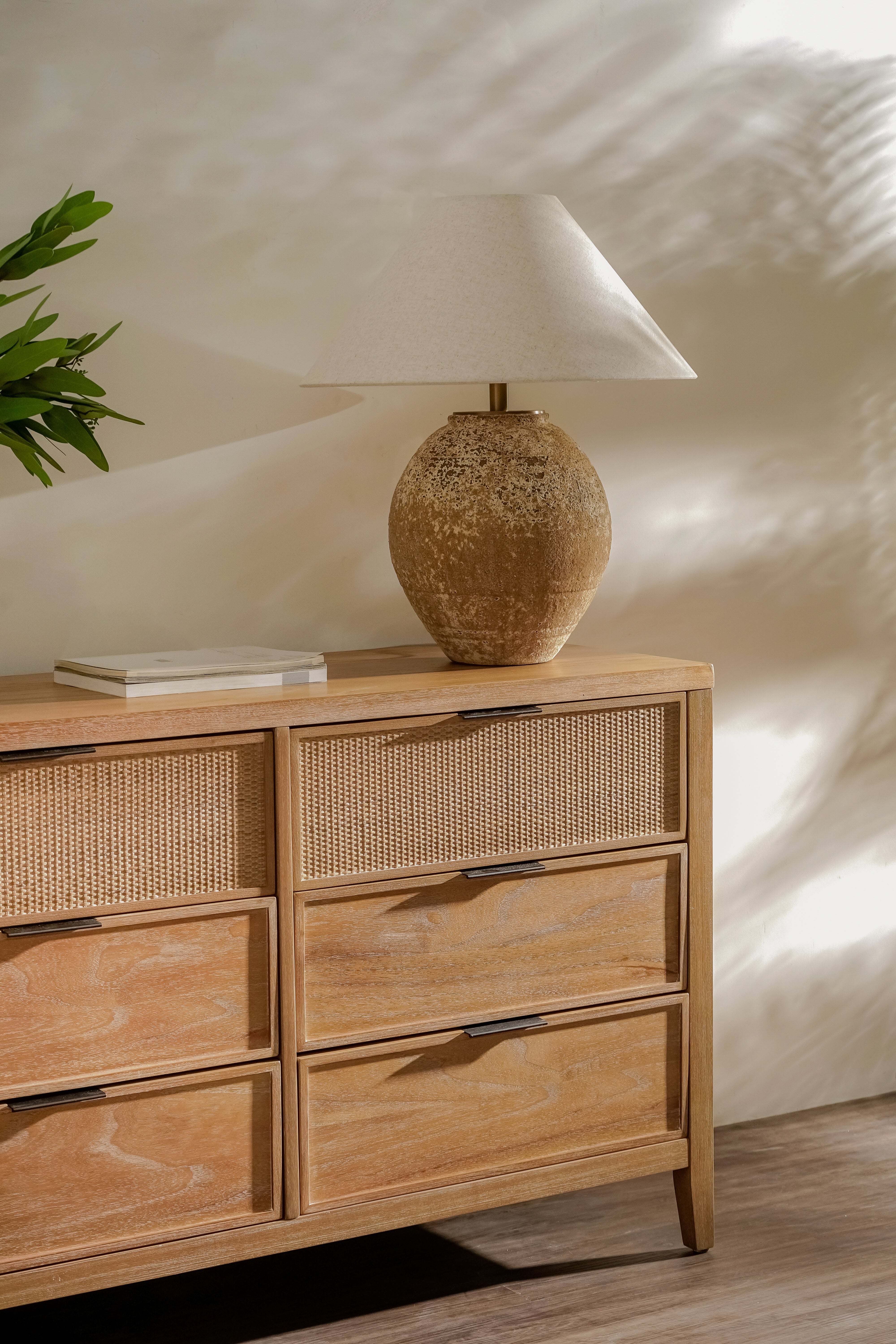 Wooden dresser with wicker drawers and a textured table lamp on top against a neutral wall.
