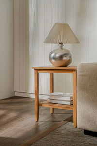 Wooden side table with a silver lamp and books against a neutral wall.