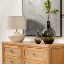 Wooden dresser with a lamp, vase, and plant against a textured wall.