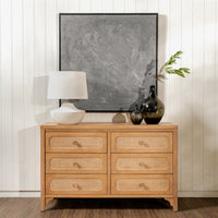 Wooden dresser with wicker drawers, lamp, and decor items against a white wall.