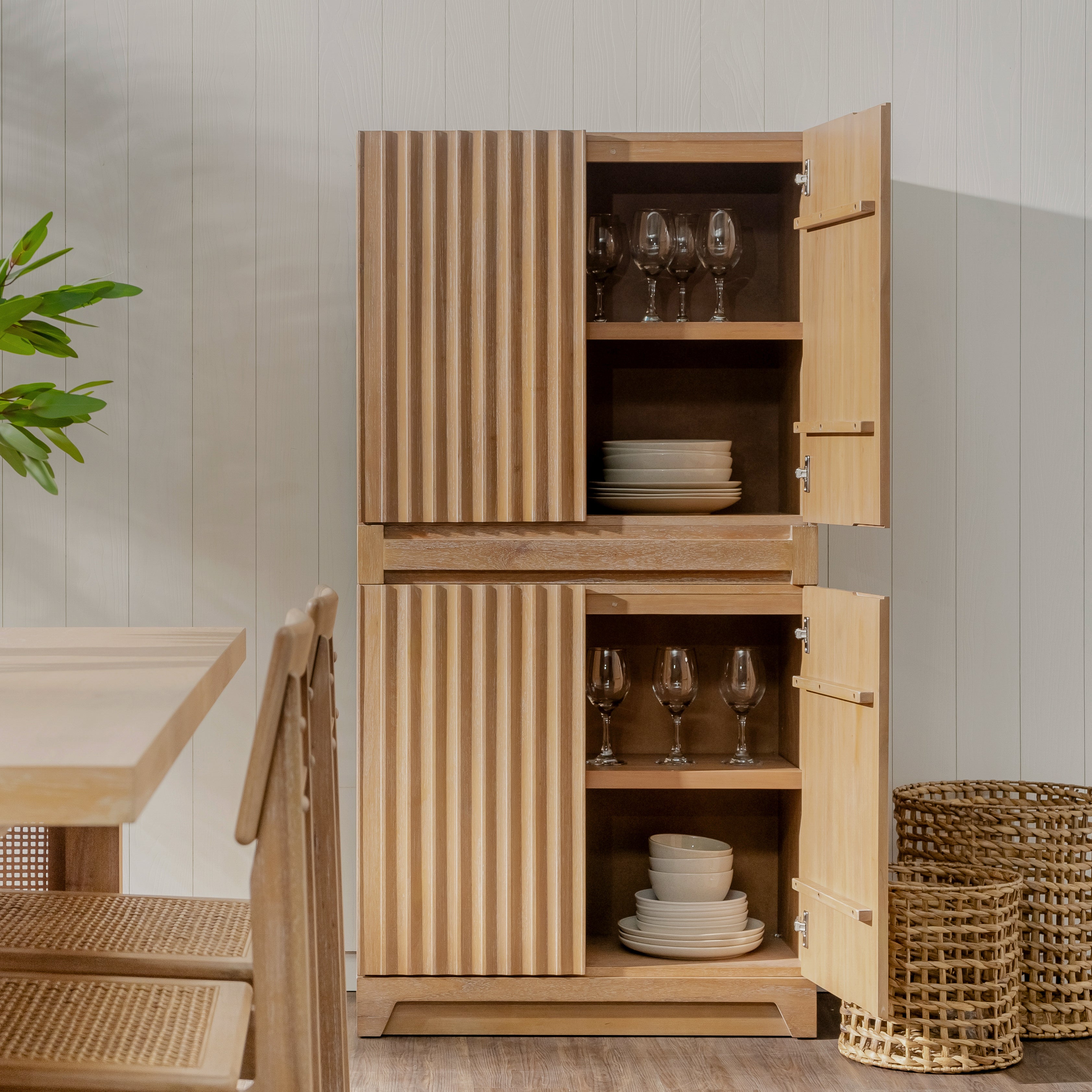 Wooden cabinet with open doors displaying glasses and plates in a room with a table and plants.
